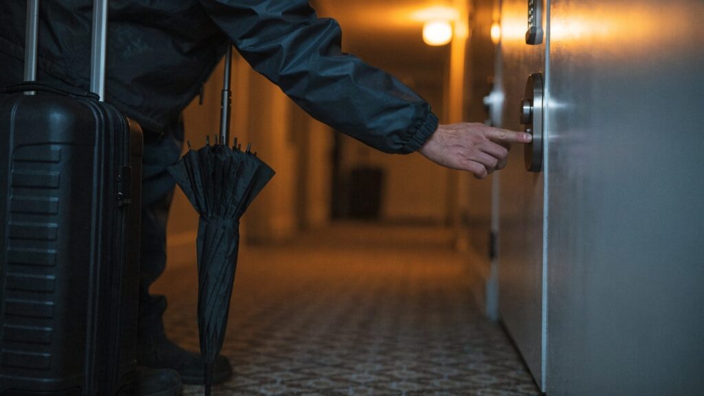 A traveler's hand pressing the lit keypad of a door lock at night, suitcase on the corridor carpet