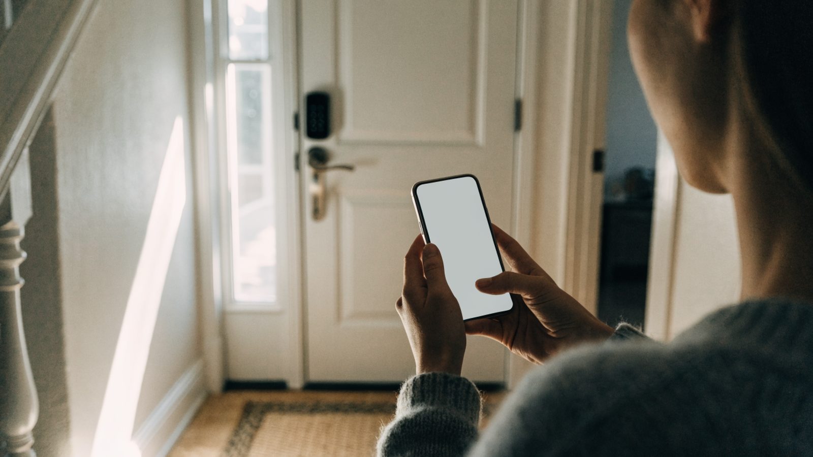 Holding a phone in a sunlit hallway facing a front door fitted with a keypad smart deadbolt