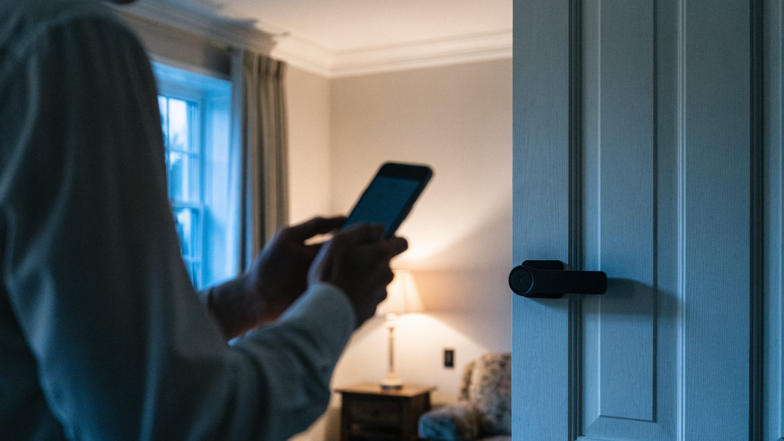 A slim smart deadbolt on a pale front door seen across a living room at dusk while someone holds a phone