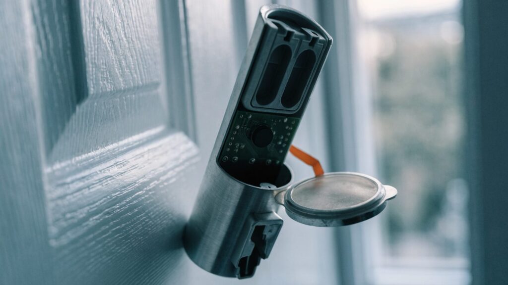 Interior deadbolt escutcheon with its cover lifted off, showing a small recessed reset button and a ribbon cable
