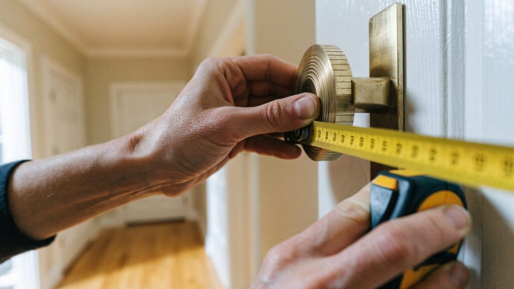 Hands holding a tape measure across the round interior rose of a brass deadbolt on a white six-panel door