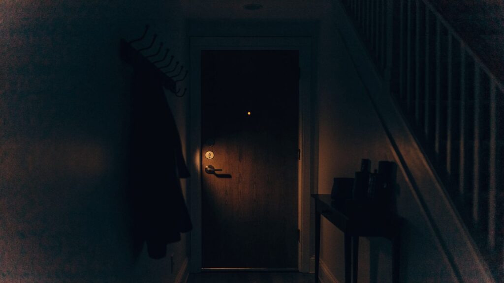 Dark entryway at night with a small amber glow from the door lock, the hallway around it in silhouette
