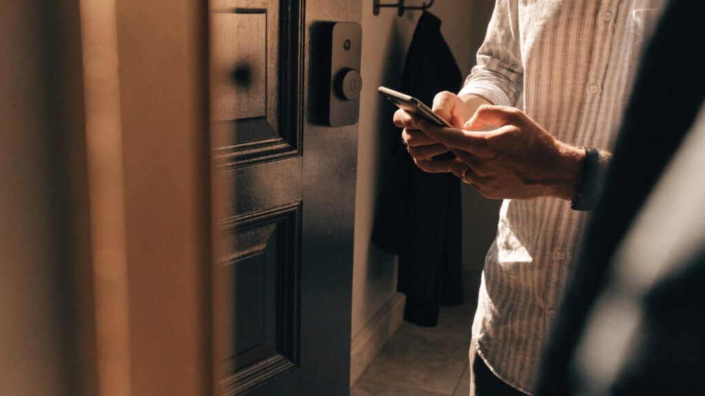 Hands holding a phone in front of a dark front door fitted with a black smart deadbolt