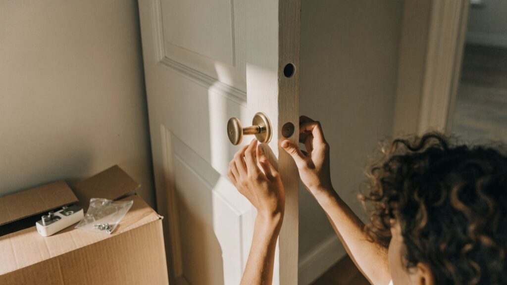 Hands refitting the original brass thumbturn to an apartment door beside a moving box
