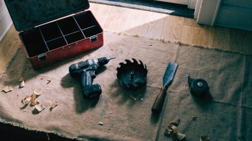 Open toolbox with a drill, hole saw, chisel and tape measure laid out on a drop cloth in a hallway