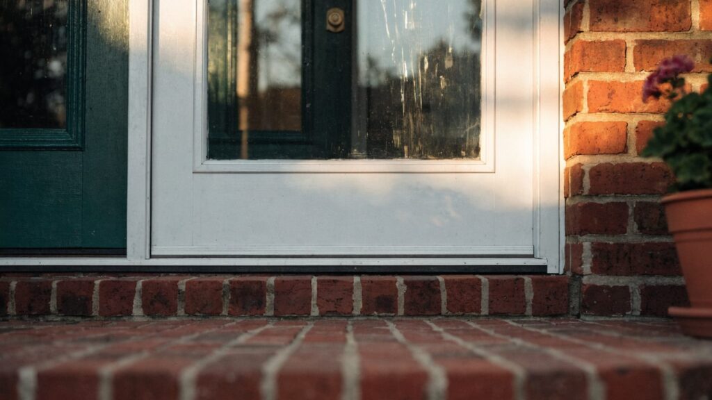 White aluminum storm door closed in front of a dark green wooden entry door on a red brick porch