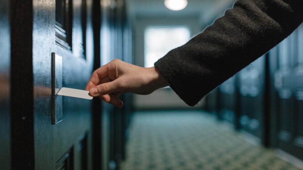 A hand holding a blank white key card beside an unmarked reader plate on a dark corridor door
