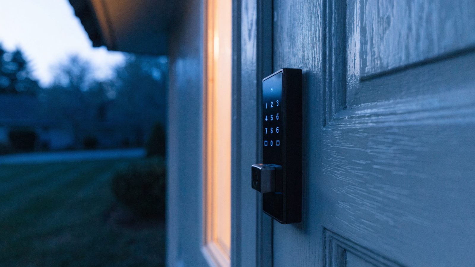 A keypad smart deadbolt glowing faintly on a gray exterior door at dusk