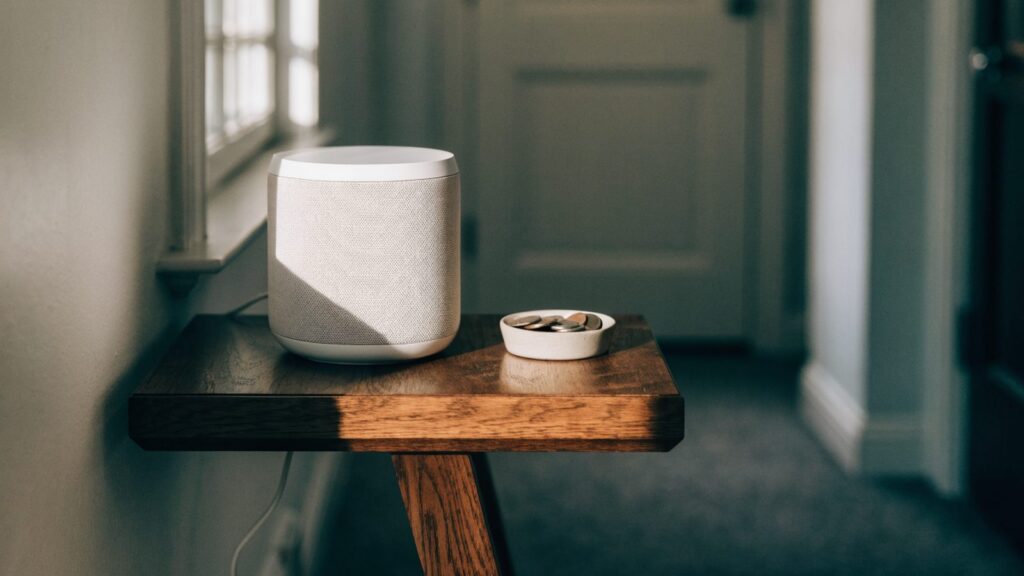 Compact matte white smart speaker on a walnut hallway console table, power cable running to a baseboard outlet