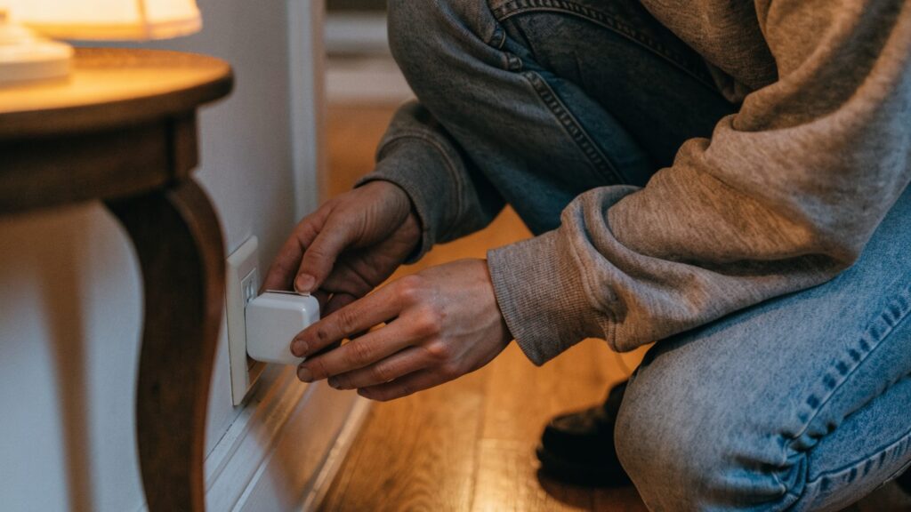 A person crouched at a baseboard outlet, both hands pressing a small white plug-in adapter into the socket