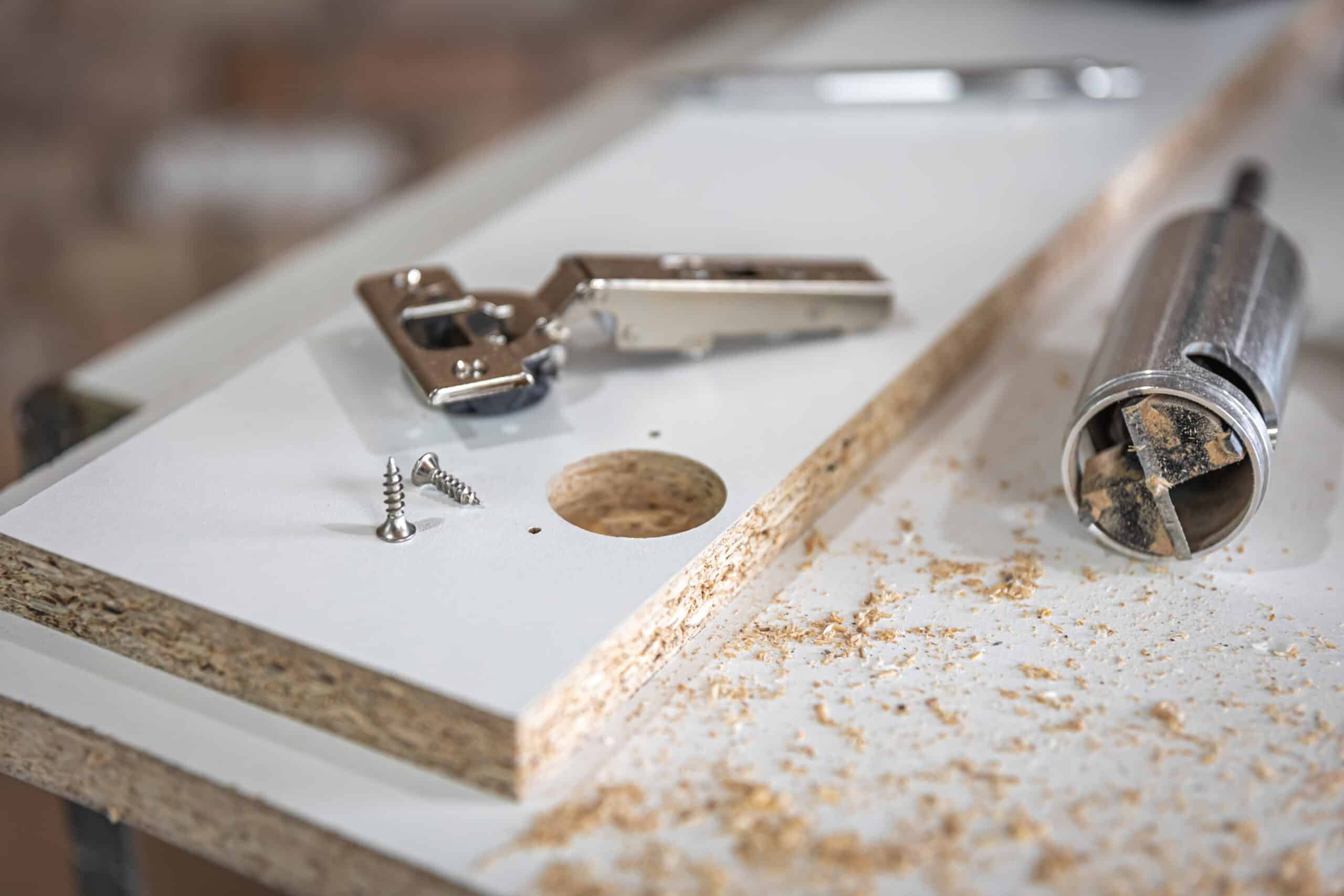 A drilled hole in a particleboard panel edge beside two wood screws, a cabinet hinge, and a hole saw bit resting in sawdust.