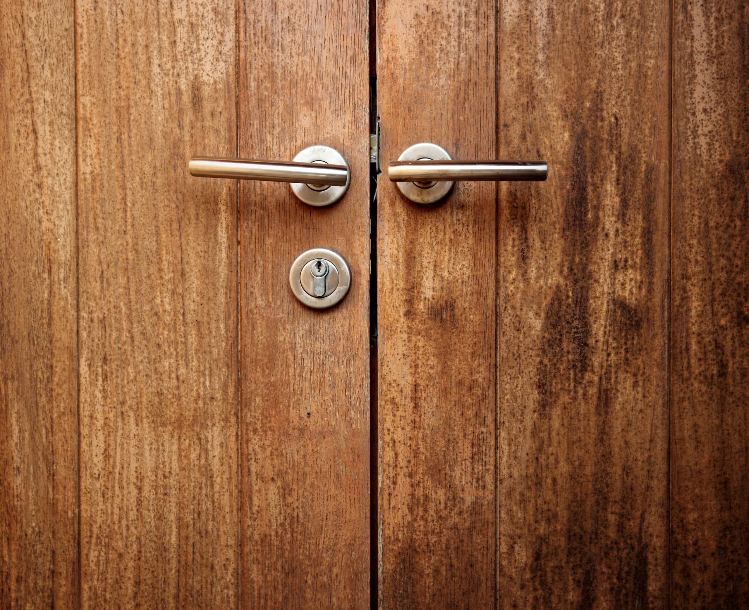 A pair of weathered wood doors fitted with lever handles and a keyed cylinder lock between them.