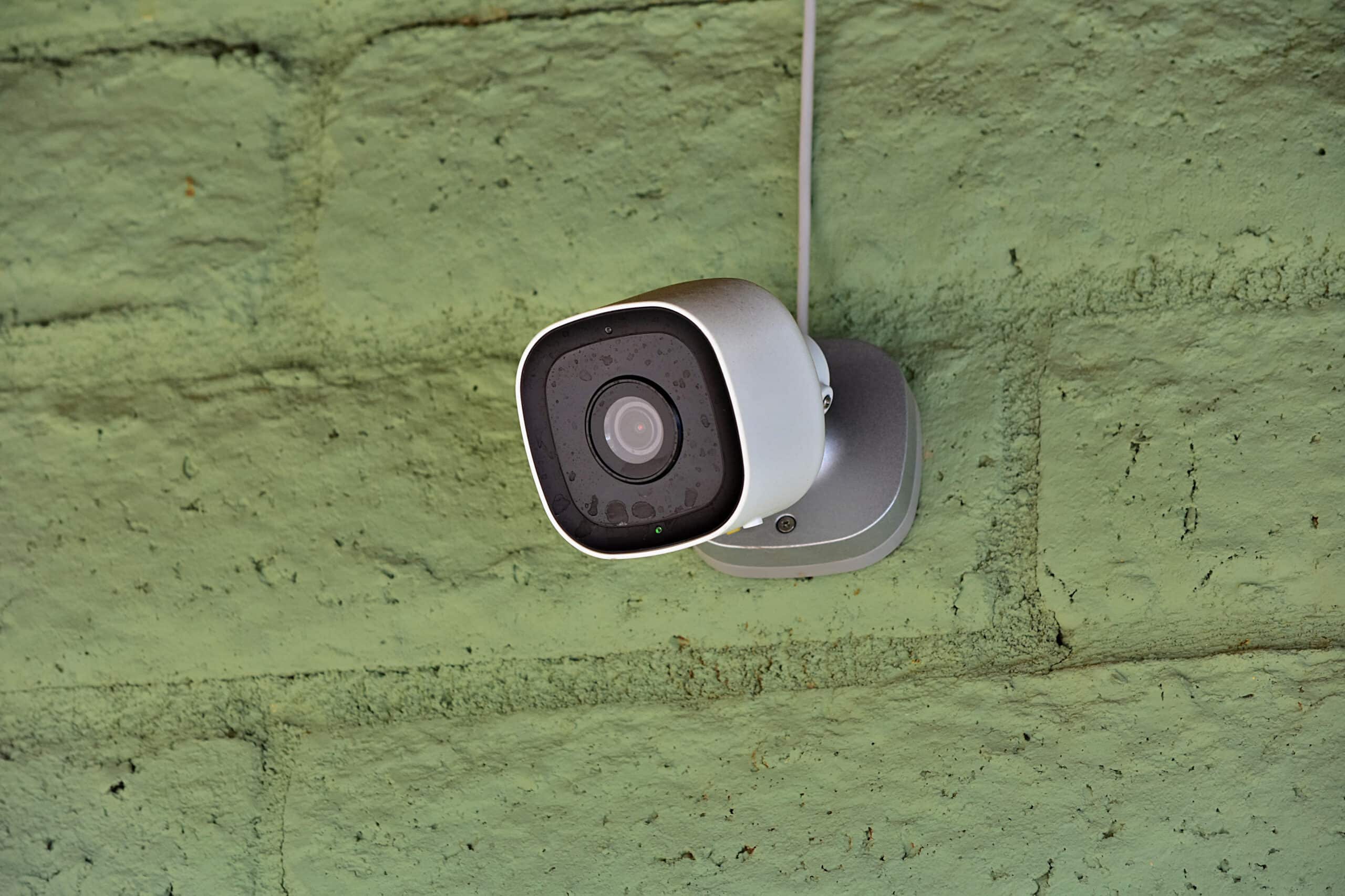 A white outdoor security camera mounted on a green painted wall, with its cable running up from the housing.